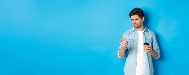 Şüpheci ve hayal kırıklığına uğramış bir adam telefonda üzücü bir mesaj okuyor, elinde kahve fincanı, mavi arka planda duruyor..