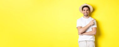 Genç erkek turist sağı işaret ediyor, gülümsüyor ve reklam veriyor, turizm ve yaşam tarzı kavramı, sarı arka plan.