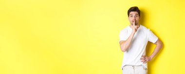 Heyecanlı bir adamın sürpriz planlaması, kameraya şşşş yapması, sessiz kalmak için parmağa basması, sarı arka planda durması..