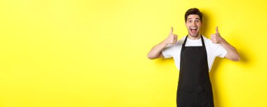 Siyah önlüklü neşeli adam başparmağını kaldırıyor, kafe ya da restoran tavsiye ediyor, sarı arka planda duruyor..