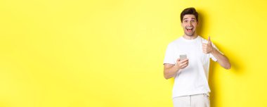 Akıllı telefonu tutan mutlu bir adam, onaylanmış bir şekilde baş parmağını gösteriyor, sarı arka planda duran, online bir şey öneriyor..