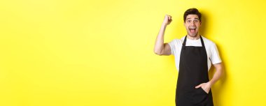 Neşeli satıcı yumruk tokuşturuyor, seviniyor ve zafer kazanıyor, sarı arka plana karşı siyah önlük içinde duruyor..