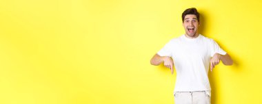 Şaşkın yakışıklı adam parmaklarını aşağı gösteriyor, pankartı heyecanla gösteriyor, sarı arka planda duruyor..