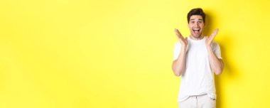 Şaşırmış ve mutlu bir adam anonslara tepki veriyor, gülümsüyor ve şaşırmış görünüyor, sarı arka planda duruyor..