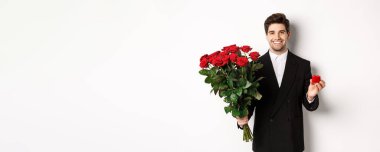 Siyah takım elbiseli yakışıklı bir adam, elinde bir buket kırmızı gül ve yüzük, evlenme teklif ediyor, kendinden emin gülümsüyor, beyaz arka planda duruyor..