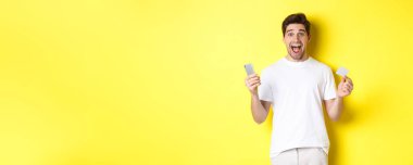 Elinde akıllı telefon ve kredi kartı olan şaşırmış bir adam. Kara Cuma 'da online alışveriş yapıyor. Sarı arka planda duruyor..