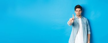 Kızgın 25 yaşındaki adam onaylamadığı için parmağını sallıyor, hayal kırıklığına uğruyor, kötü bir şeyi yasaklıyor, hayır diyor, mavi arka planda duruyor..