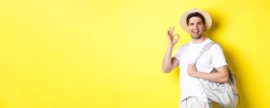 Turizm, seyahat ve tatil konsepti. Mutlu bir turist tatile çıkıyor, sırt çantası taşıyor ve tamam işareti gösteriyor. Gülümsüyor, sarı arka planda duruyor..