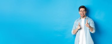 Kendine güvenen bir adam tebrik ediyor, göz kırpıyor ve seni işaret ediyor. Mavi arka planda mutlu bir şekilde duruyor ve gülümsüyor..