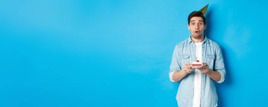 Parti konisinde elinde doğum günü pastası olan yakışıklı bir adam, şaşırmış görünüyor, mavi arka planda duruyor..