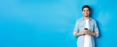 Kahve dükkanından kahve içen, gülümseyen, mavi arka planda duran mutlu genç adam..