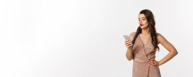 Noel partisi ve kutlama konsepti. Moda elbisesi giymiş dikkatsiz bir kadın cep telefonundan mesaj okuyor, şüpheli görünüyor, beyaz arka planda duruyor..