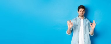 Tatmin olmuş yakışıklı bir adam iyi bir izlenim veriyor ve memnun görünüyor, iyi bir şeyi onaylıyor, mavi arka plana karşı duruyor..