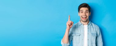 Yakışıklı sakallı adamın yakın çekimi gülümsüyor, bir numaralı parmağını gösteriyor, mavi arka planda duruyor..