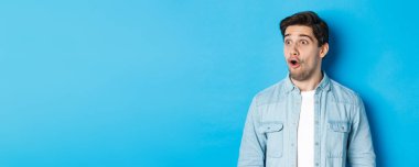 Şaşırmış, soldan etkilenmiş, ağzı açık, büyüleyici, sıradan kıyafetler giymiş, mavi arka planda duran yakışıklı adamın yakın çekimi..