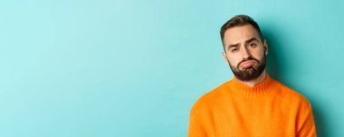 Üzgün, hayal kırıklığına uğramış ve sıkıntılı görünen, turkuaz arka planda turuncu süveter giyen üzgün genç adamın yakın çekimi..