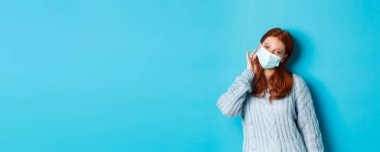 Kış, covid-19 ve sosyal uzaklık kavramı. Güzel kızıl saçlı genç kız, yüz maskesi takıyor ve saçlarını kulaklarının arkasına sıkıştırıyor, kameraya bakıyor, mavi arka planda duruyor..