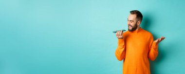 Hoparlörden konuşan mutlu bir adam, cep telefonuna işaret edip sesli mesaj kaydediyor, açık mavi arka planda turuncu süveter giyiyor..