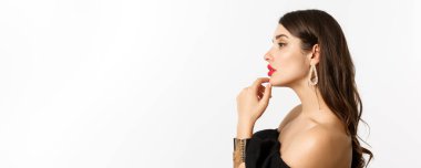 Moda ve güzellik konsepti. Siyah gece elbisesi, makyaj ve küpe giymiş, soldan şehvetli görünen, beyaz arka planda duran çekici ve şık bir kadın profili..