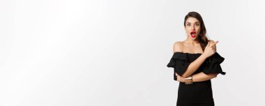 Moda ve güzellik. Siyah elbiseli güzel bir kadın, kırmızı dudaklı, reklam teklifini işaret eden, kameraya bakan, beyaz arka planlı,.