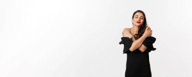 Moda ve güzellik. Siyah elbiseli, şehvetli ve güzel bir kadın, kapalı gözlerle kendine sarılıyor, hayal kuruyor, beyaz arka planda duruyor..