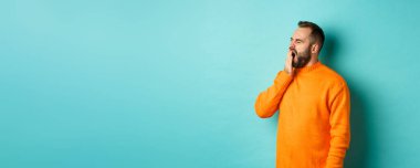 Yorgun sakallı bir adamın esneme fotoğrafı, ağzı elle kaplanmış, açık mavi arkaplana karşı uykulu duruyor..