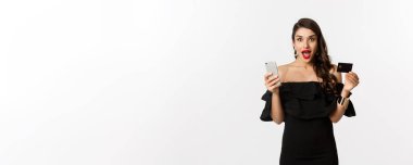 İnternetten alışveriş konsepti. Siyah elbiseli şık bir kadın, elinde akıllı telefonlu bir kredi kartı var, heyecanlı görünüyor, beyaz arka planda duruyor..