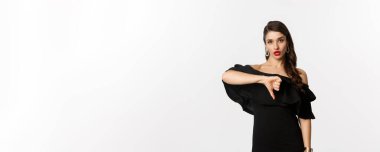 Moda ve güzellik. Siyah elbiseli, hayal kırıklığına uğramış, baş parmağını aşağı gösteren, kötü bir şeyden hoşlanmayan, beyaz arka plana bakarak yargılayan, küstah bir kadın..