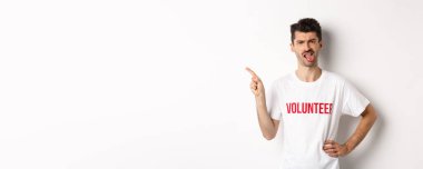 Hayal kırıklığına uğramış gönüllüler kötü reklam teklifleri sunuyor, dil yapıştırıyor ve iğreniyor, parmağını sola gösteriyor, örgüt tişörtü giyiyor, beyaz arka plan.