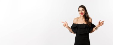 Moda ve güzellik. Mücevher, makyaj ve siyah elbise giymiş çekici bir kadın, gülümseyen ve parmaklarını yana doğru gösteren fotokopi alanı teklifi, beyaz arka plan.