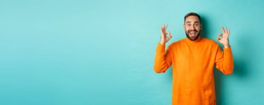 Etkileyici bir adam harika bir şeyi övüyor, iyi işaretler gösteriyor ve şaşırmış görünüyor, açık mavi arka planda turuncu süveter içinde duruyor..