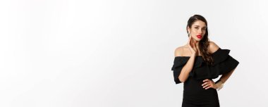 Siyah elbiseli şehvetli genç bir kadın, küpelerini gösteriyor ve kameraya seksi bakıyor, beyaz arka planda duruyor..