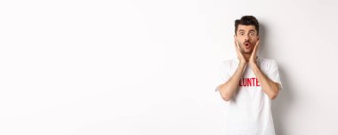 Gönüllü tişörtlü şok olmuş bir adam, hayranlıkla kameraya bakıyor, beyaz arka planda duruyor..