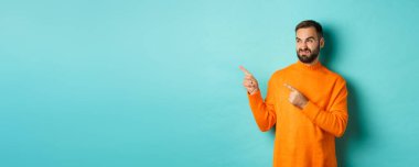 Tatminsiz ve şüpheci adam somurtuyor, kötü ürün gösteriyor, hayal kırıklığıyla sol parmaklarını gösteriyor, turkuaz arka planda duruyor..