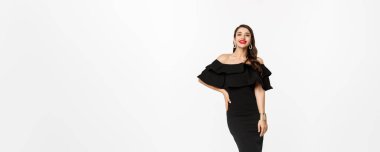 Güzellik ve moda konsepti. Tam boy, güzel esmer kadın, lüks siyah elbiseler ve topuklu ayakkabılar giyiyor, kırmızı dudaklarıyla gülümsüyor, beyaz arka planda dikiliyor..