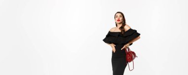 Güzellik ve moda konsepti. Siyah elbiseli, topuklu ayakkabılı, kendine güvenen, kameraya küstah ve beyaz arka planlı zarif bir genç kadın..