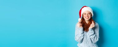 Kış tatili ve Noel arifesi konsepti. Noel Baba şapkalı umut dolu kızıl kız, Noel Baba 'dan dilek diliyor, dua ediyor, Noel Baba şapkası takıyor, mavi arka planda duruyor..