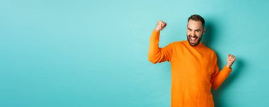 Kazandım. Heyecanlı adam zafer kazanıyor, yumruk tokuşturuyor ve evet diyor, zafer kazanıyor ve tatmin olmuş görünüyor, hedefe ulaşıyor, açık mavi arka planda duruyor..