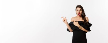 Moda ve güzellik. Siyah cazibeli bir elbise giymiş, solu işaret eden, reklamını yapan ve beyaz arka planda şaşkın şaşkın bakan şaşırmış bir kadın..