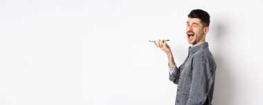 Mutlu adam kameraya bakarken ses çevirici uygulamasını kullanıyor ya da hoparlörden konuşuyor, telefonu ağıza yakın tutuyor, beyaz arka planda duruyor..