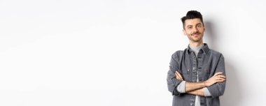 Gururlu yakışıklı adam tatmin olmuş görünüyor, göğsünde çapraz kollar ve kameraya gülümsüyor, iyi bir şeye bakıyor, beyaz arka planda duruyor..