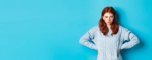 Redhead girl staring angry and displeased at camera, frowning upset, standing against blue background.