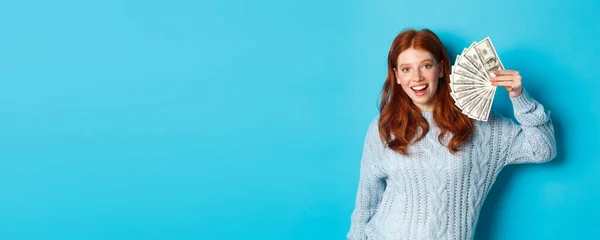 Cheerful redhead woman in sweater showing dollars, smiling pleased and holding money, standing over blue background.