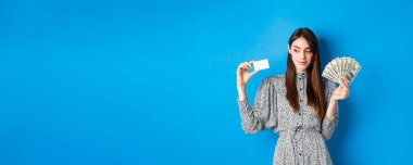Alışverişe. Elbiseli güzel kız yana bakıyor ve gülümsüyor dalgın dalgın, para ve kredi kartı gösteriyor, mavi arka planda duruyor..