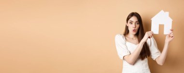Gayrimenkul. Heyecanlı bir kadın gazete kupürlerini gösterip hayret verici bir yüz ifadesi takınıyor, ev alıyor, bej arka planda duruyor..