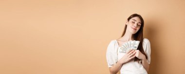 Zarif bir kız, mutlu bir gülümsemeyle ve kapalı gözlerle parayı kucaklıyor, dolar banknotları gibi, bej arka planda duruyor..