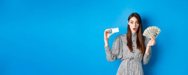 Heyecanlı şanslı kız plastik kredi kartı ve para gösteriyor, hayret içinde, pahalı bir şey alıyor, mavi arka planda duruyor..