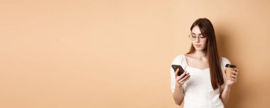 Gözlüklü kız cep telefonundan mesaj okuyor, kahve içiyor, bej arka planda duruyor..