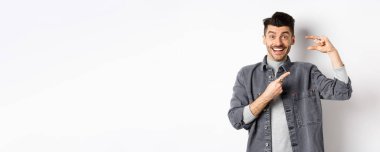 Heyecanlı adam küçük boy gösteriyor, küçük bir şeyi işaret ediyor ve gülümsüyor, beyaz arka planda duruyor..