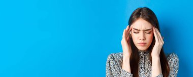 Genç bir kadının yakın plan portresi hasta, baş şakaklarına dokunuyor ve baş ağrısından çatık kaşlarını çatıyor, migren oluyor, mavi arka planda duruyor..